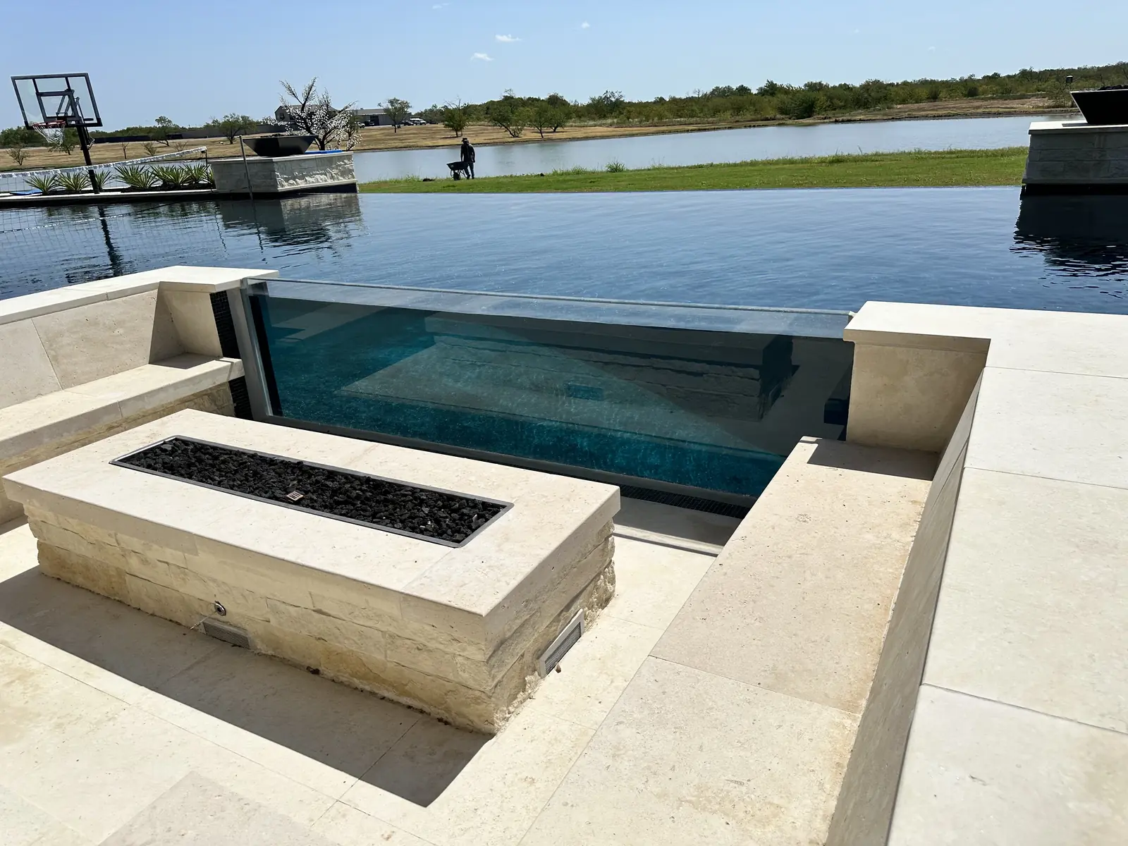 A glass-walled infinity pool at a lakefront property in Southlake, Texas, with a stone fire pit on the deck and the pool's water visible through an acrylic panel set into its wall