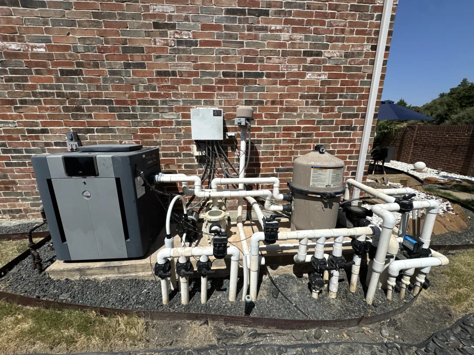 A pool equipment pad against a brick wall: a heater, a cartridge filter, pumps and an automation panel, connected by white PVC plumbing with valves