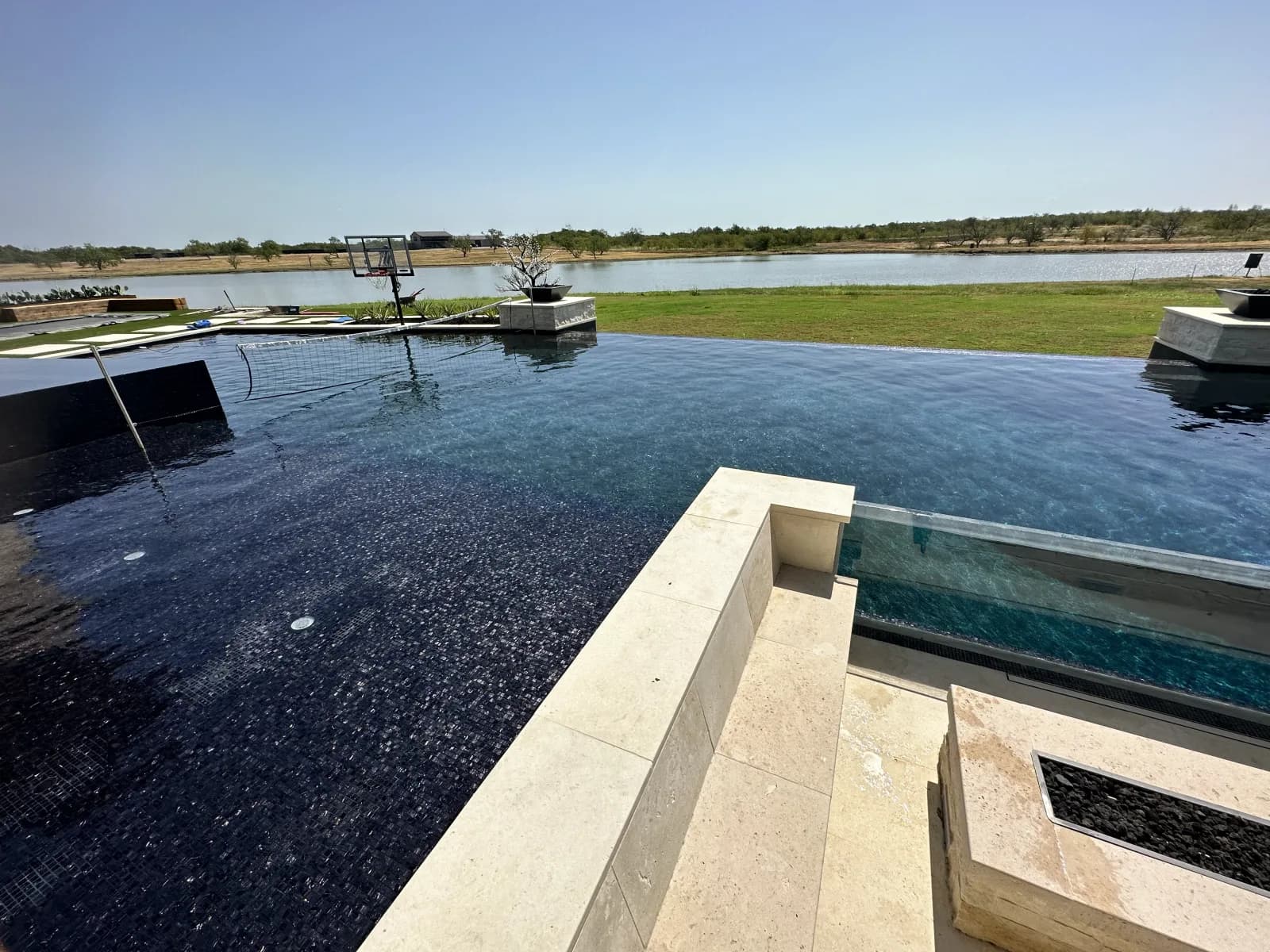 A dark-bottomed infinity pool looking out over a lake, with a stone fire feature on the deck in the foreground
