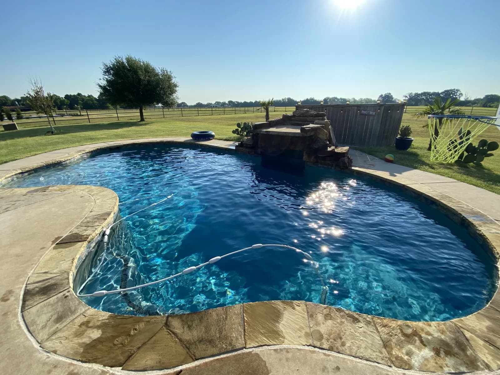 A freeform pool with clear blue water and deck jets arcing across it, a raised stone water feature behind, on a bright day